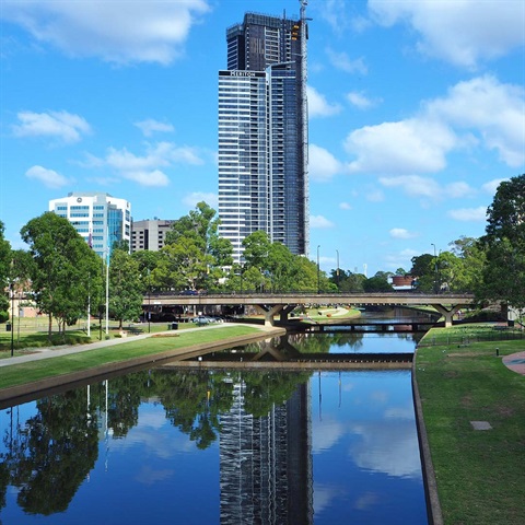 A tall skyscraper towers over a serene river, reflecting its image in the water. A bridge crosses the river, framed by green trees and a vivid blue sky.