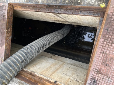 A large corrugated pipe extends into an open, rectangular manhole filled with murky water. The rusty metal frame and concrete surroundings indicate an industrial setting.