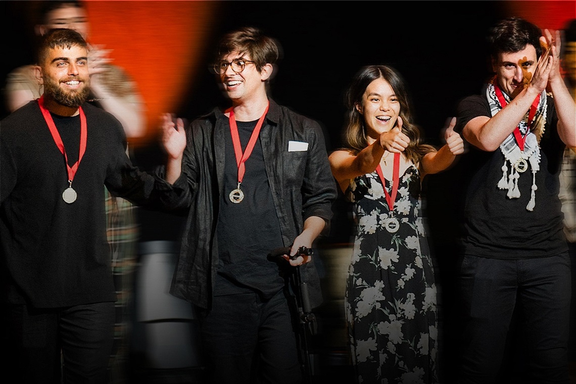 Four people stand on stage, wearing medals and smiling. They are clapping and giving thumbs up, exuding a celebratory and joyful atmosphere.