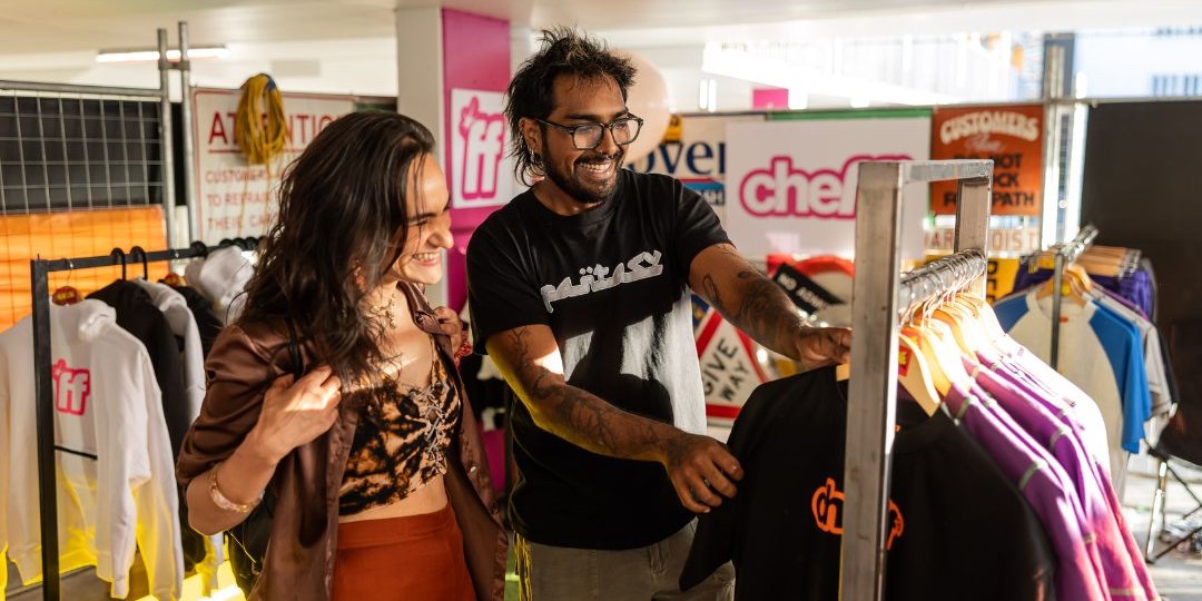 A couple smiles while browsing clothes in a cosy second-hand shop. Racks of colourful garments surround them, creating a cheerful and relaxed atmosphere.