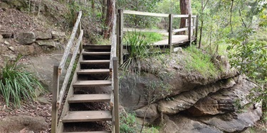 Wooden stairs with railing lead up a rocky path to a small wooden platform nestled among trees and greenery, conveying a rustic, natural feel.