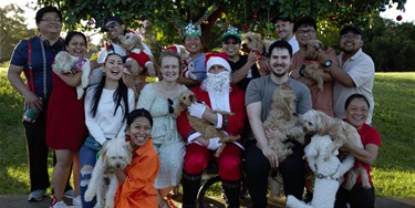 A group of cheerful people and dogs pose under a decorated tree, with one person dressed as Santa. Everyone is smiling, creating a festive, joyful atmosphere.