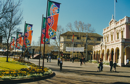 Historical image in colour showing a side view of the town hall and the park in in front, with people in the street