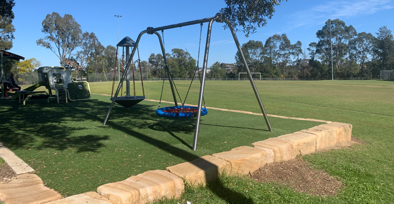 Swings and play equipment covered by tree shade, sports field to right