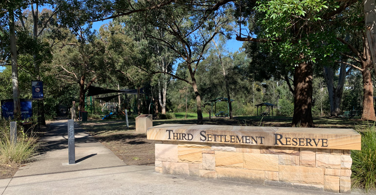 Third Settlement Reserve wall sign on right with walking path on left, play space in the background surrounded by trees