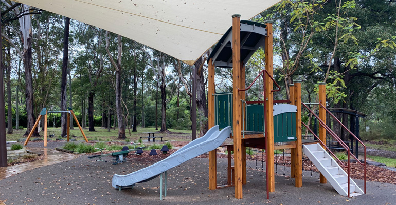 Play equipment with stairs, slide and platforms, see-saw and balancing rocks, swings and picnic table in background