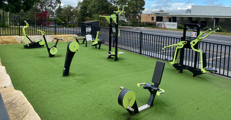 Small park with outdoor exercise gym equipment, metal bar fence and car park in background