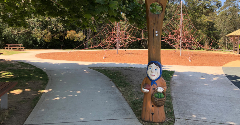 Tree with wooden statue of old woman with scarf holding an apple and a basket, two footpaths and play equipment in background