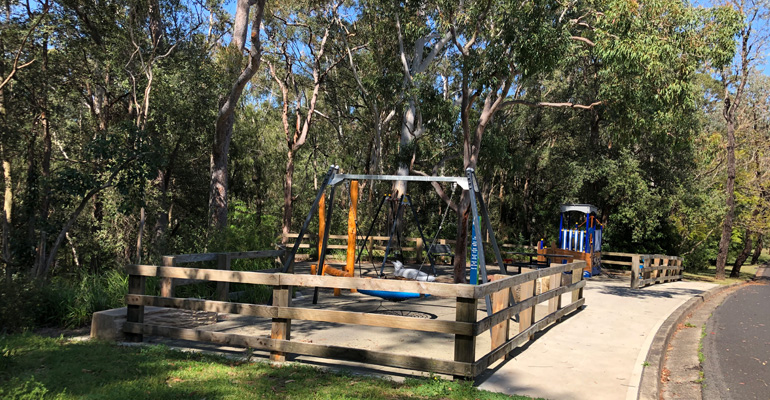 Playground surrounded by low wooden fence and trees, road to the right