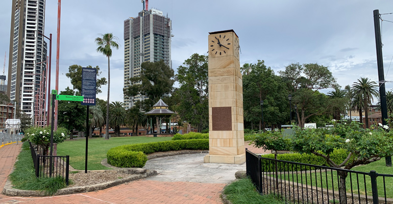 The Gollan Memorial Clock Tower made of sandstone, surrounded by hedges, high rise buildings on left 