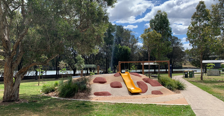 Hilled playspace with slide, swings, picnic area, trees and water in background