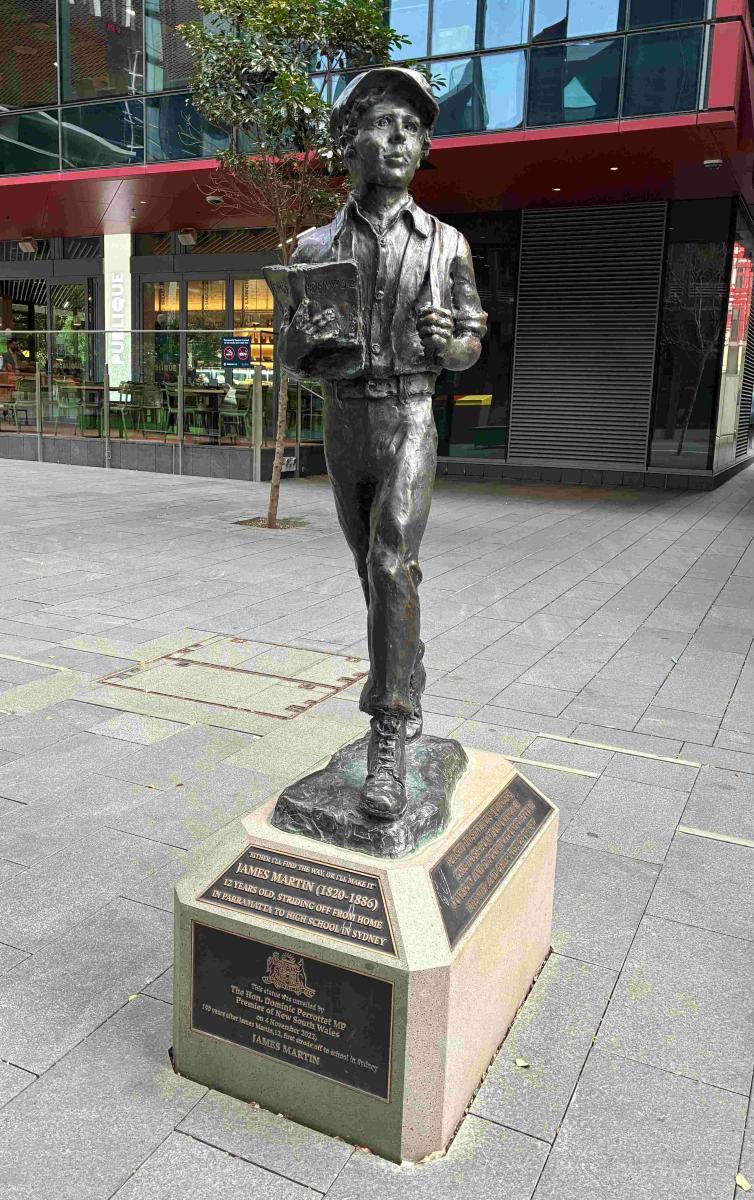 Bronze statue of a young boy in school attire holding books, mounted on a textured stone base with plaques. Located in a plaza near a modern building.