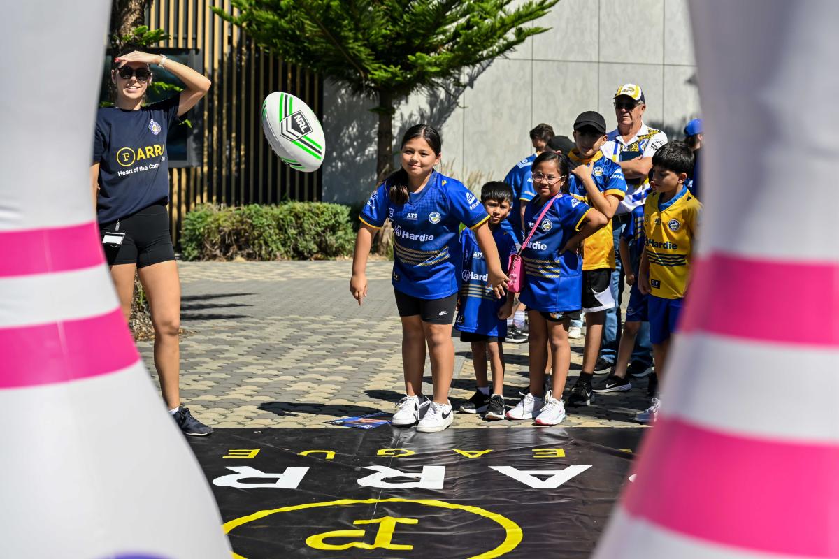 A young girl in a blue sports jersey throws a green-striped rugby ball in a playful outdoor setting, surrounded by children and a smiling adult.