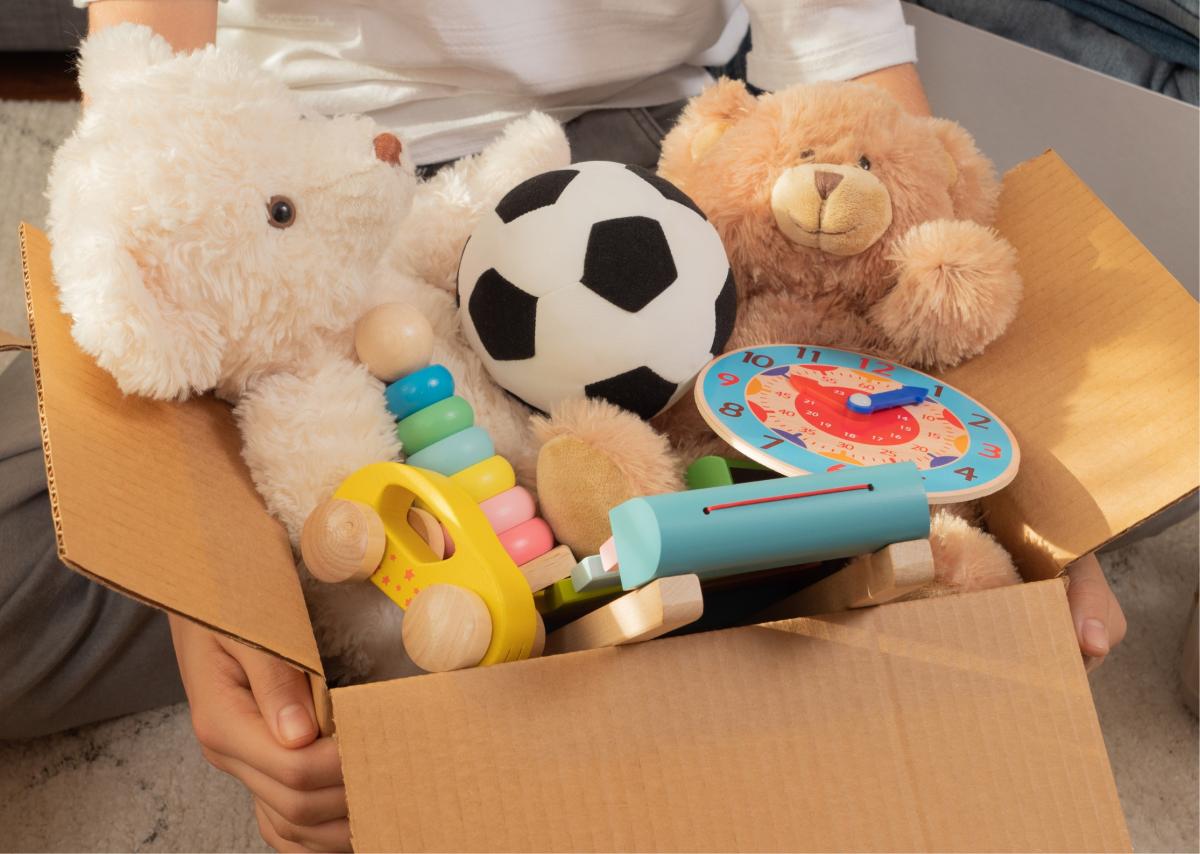 A cardboard box filled with toys, including two teddy bears, a soccer ball, a colorful ring stacker, a toy clock, and a wooden whistle. Bright and playful.
