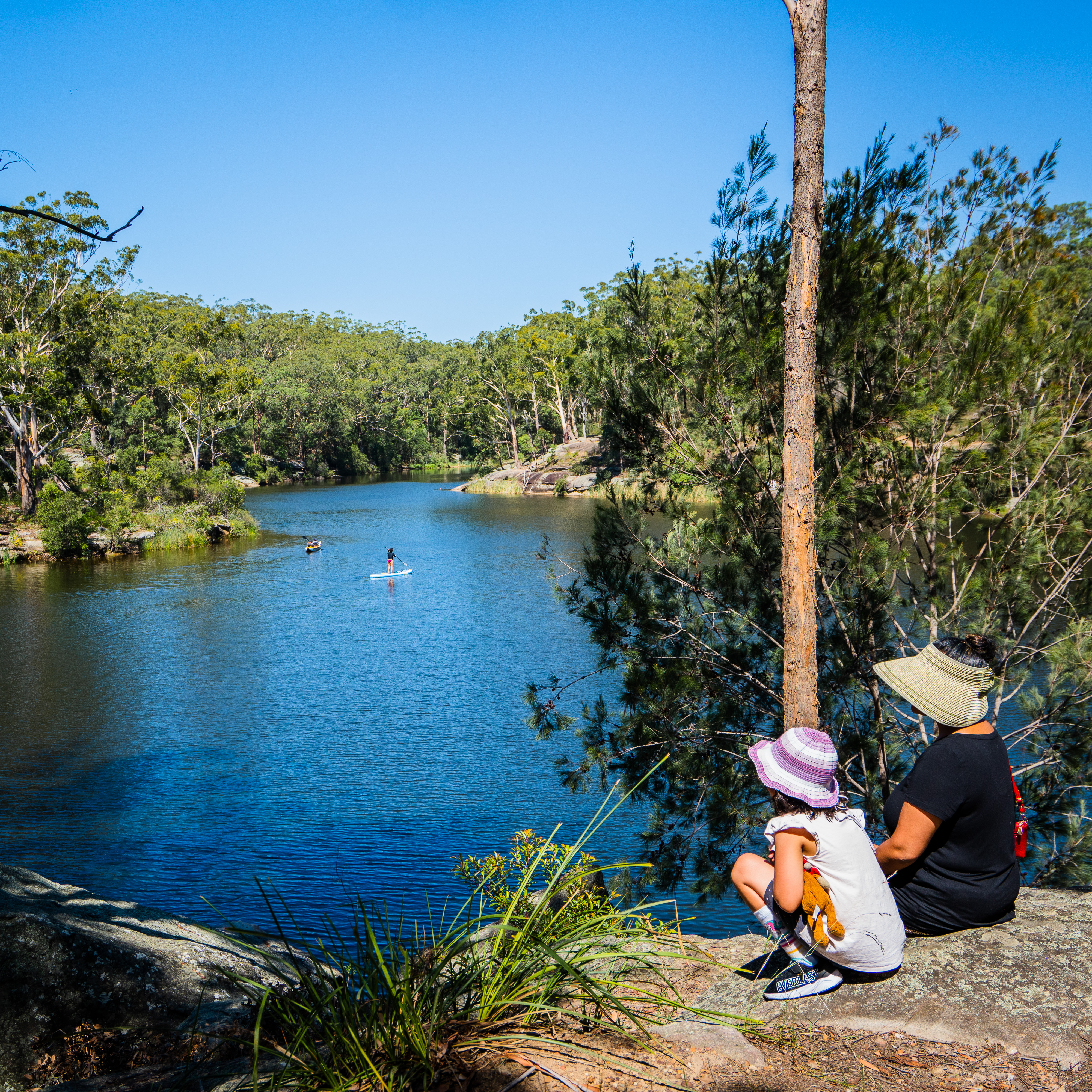 a mother and daughter sitting on a rock overlooking a lake, surrounded by green foliage