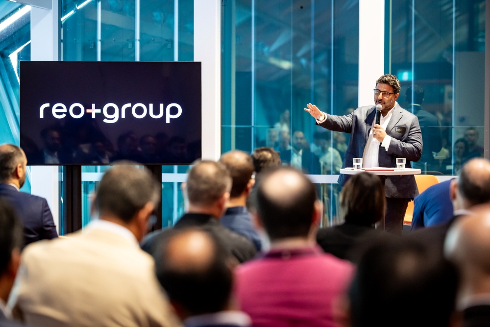 People sitting in chairs looking at man with microphone and tv screen with Reo Group logo