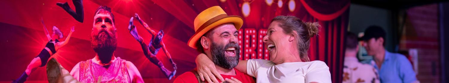 A man in a yellow hat and a woman in a patterned skirt are laughing as he lifts her in front of a circus-themed backdrop.