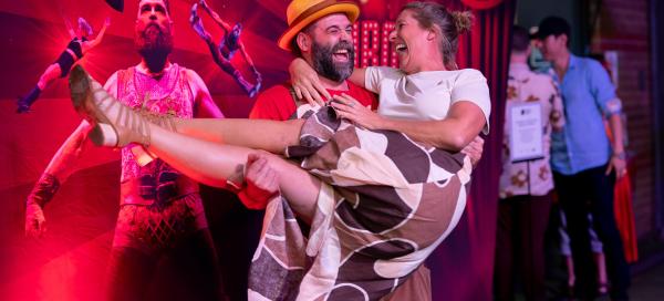 A man in a yellow hat and a woman in a patterned skirt are laughing as he lifts her in front of a circus-themed backdrop.
